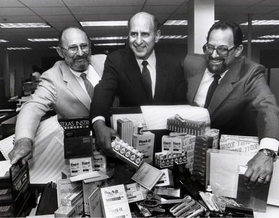 Three men smiling behind a display of office supplies in the original Quill location, 1956